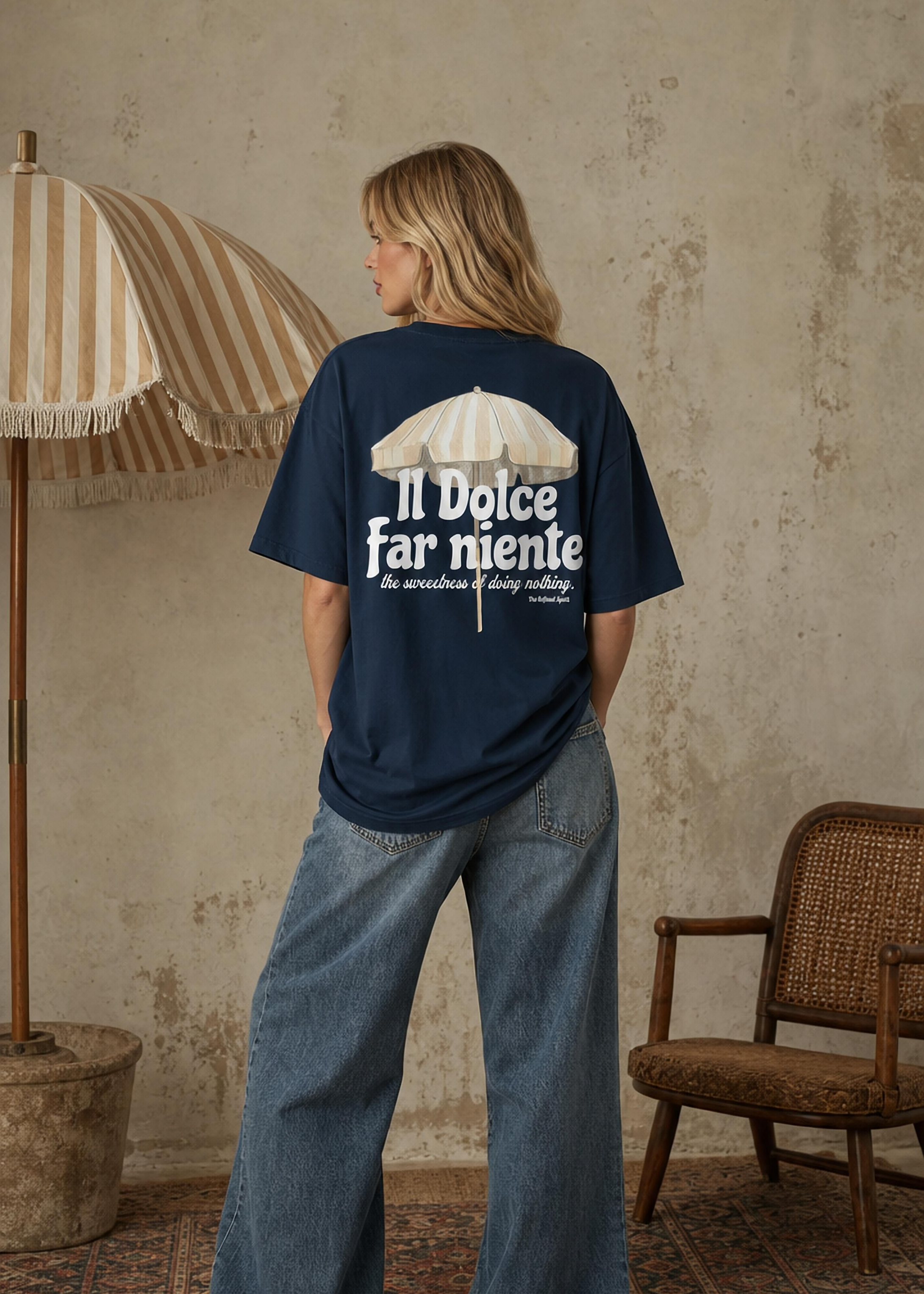 Person wearing a navy blue t-shirt with text and graphic, standing in a room with a textured wall, chair, and umbrella.