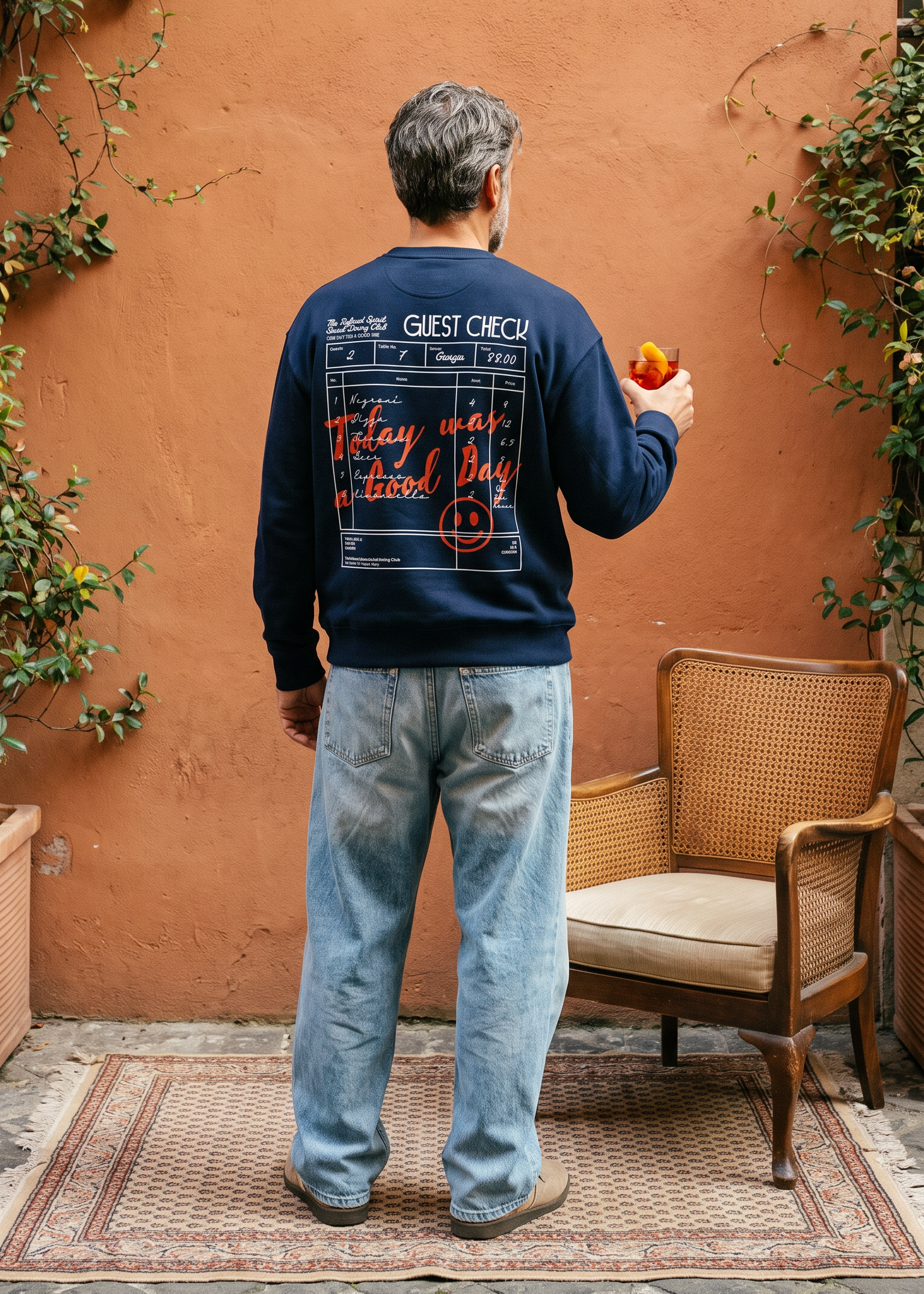 A man stands with his back to the camera in a rustic courtyard, wearing a navy blue crewneck sweatshirt with a large graphic print on the back, light blue jeans, and holding an apple. The background features a textured terracotta wall, climbing green vines, and a woven armchair.