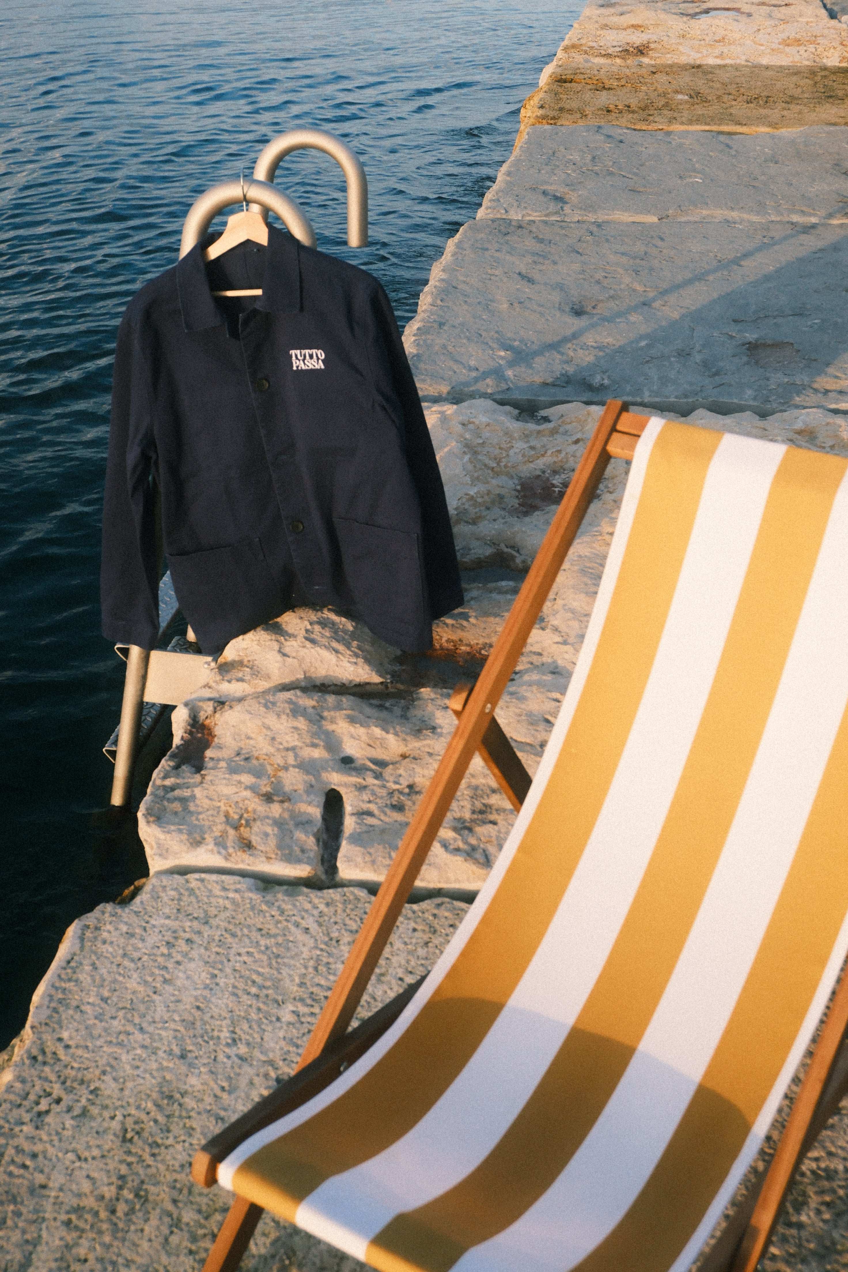 Black jacket and gold and white striped towel on a wooden chair by a rocky shoreline.