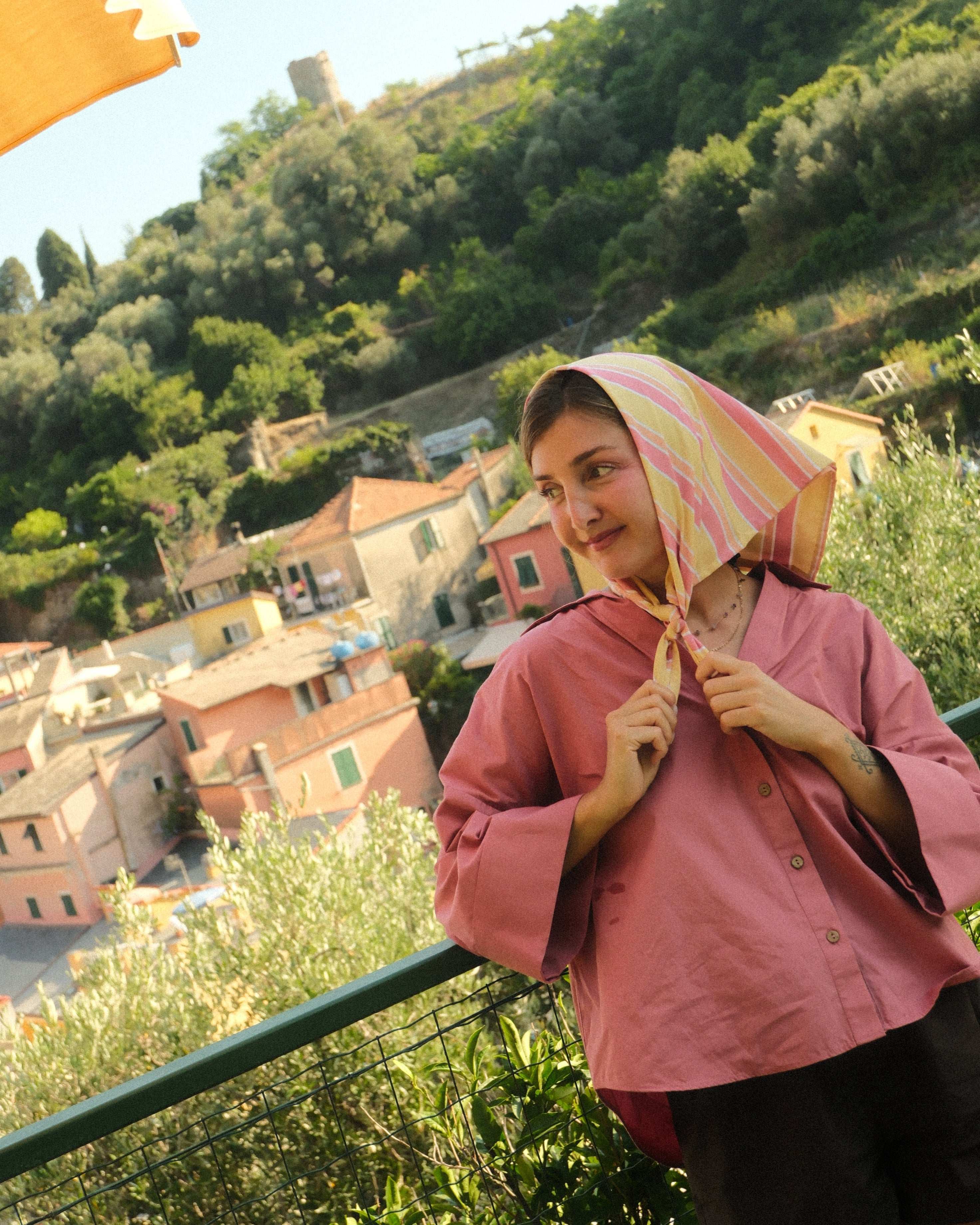 Person with a pink jacket and yellow headscarf standing on a scenic overlook with greenery and buildings in the background.