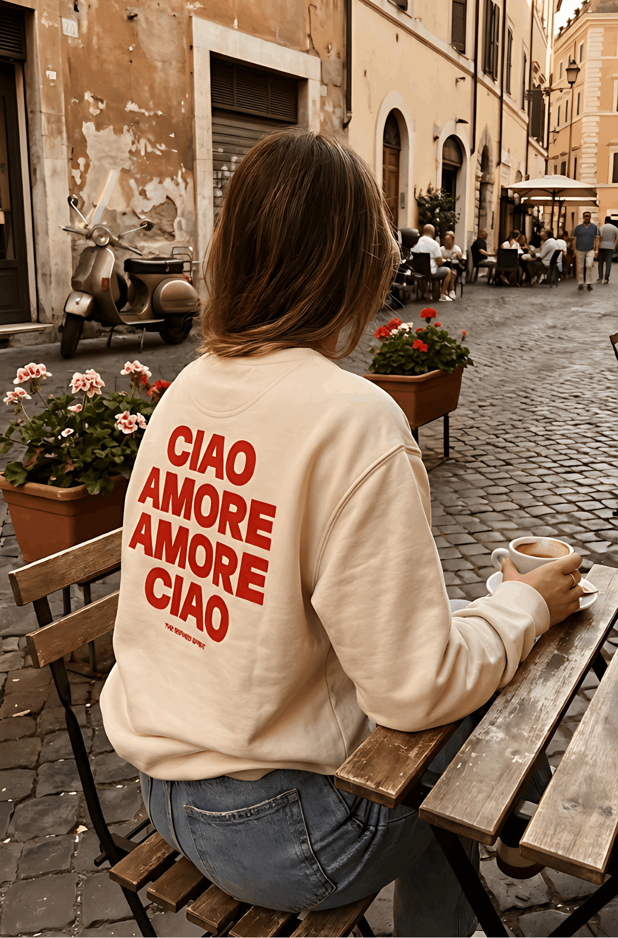 Person sitting at a table on a cobblestone street wearing a sweatshirt with 'CIAO AMORE AMORE CIAO' text.