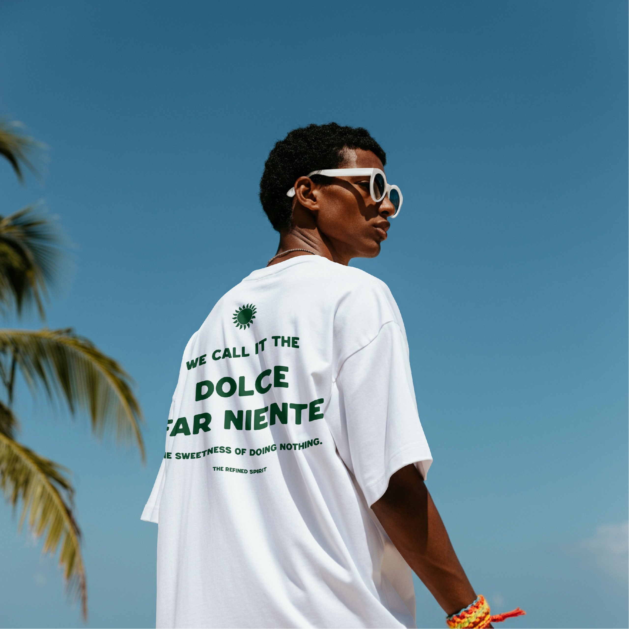 Person wearing a white t-shirt with text, standing against a clear blue sky with palm trees.