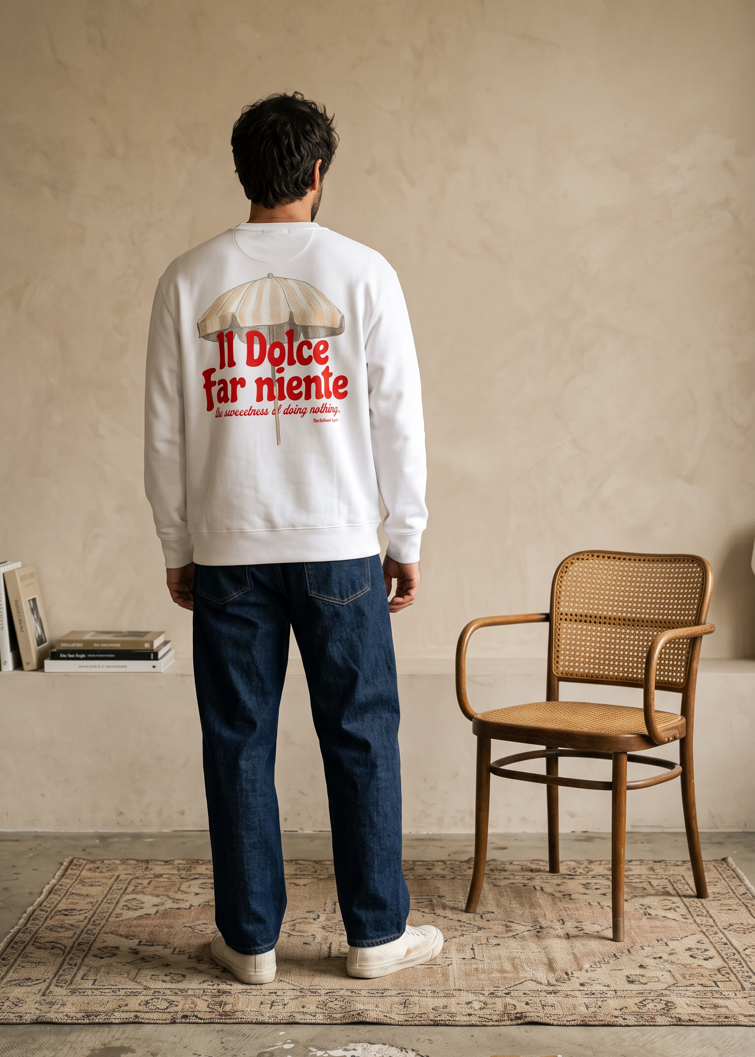A man stands with his back to the camera wearing a white sweatshirt with a red and beige graphic on the back that reads 'Il Dolce far niente'. He is wearing blue jeans and white sneakers in a minimalist room with a textured beige wall, a wooden chair, and a patterned rug.
