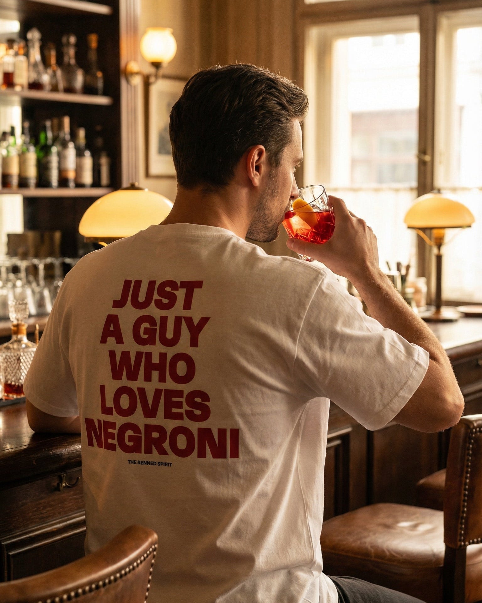 Man sitting at a bar drinking a Negroni, wearing a white t-shirt with red text.
