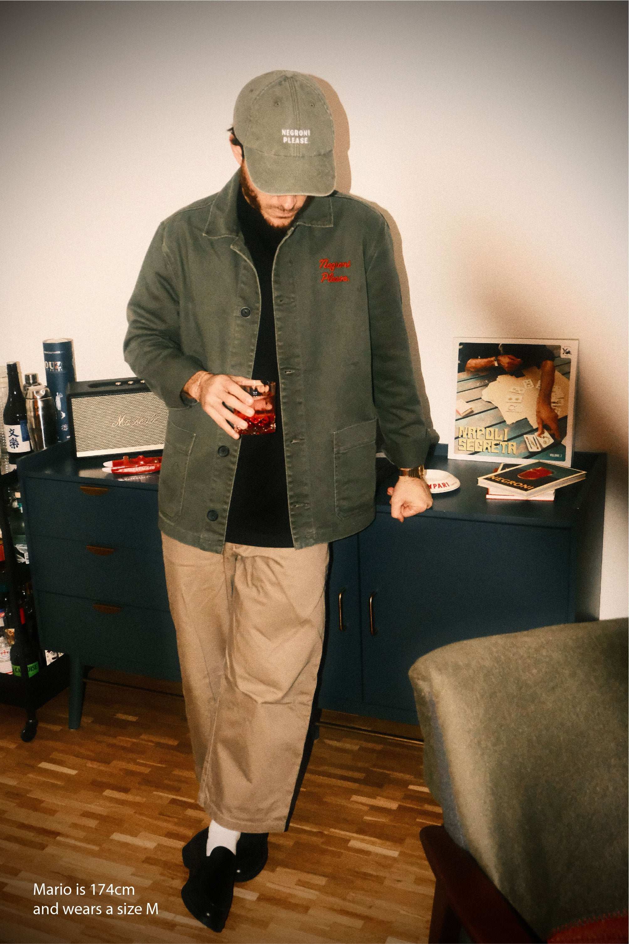 Person wearing a green jacket and beige pants standing in a room with a desk and books.