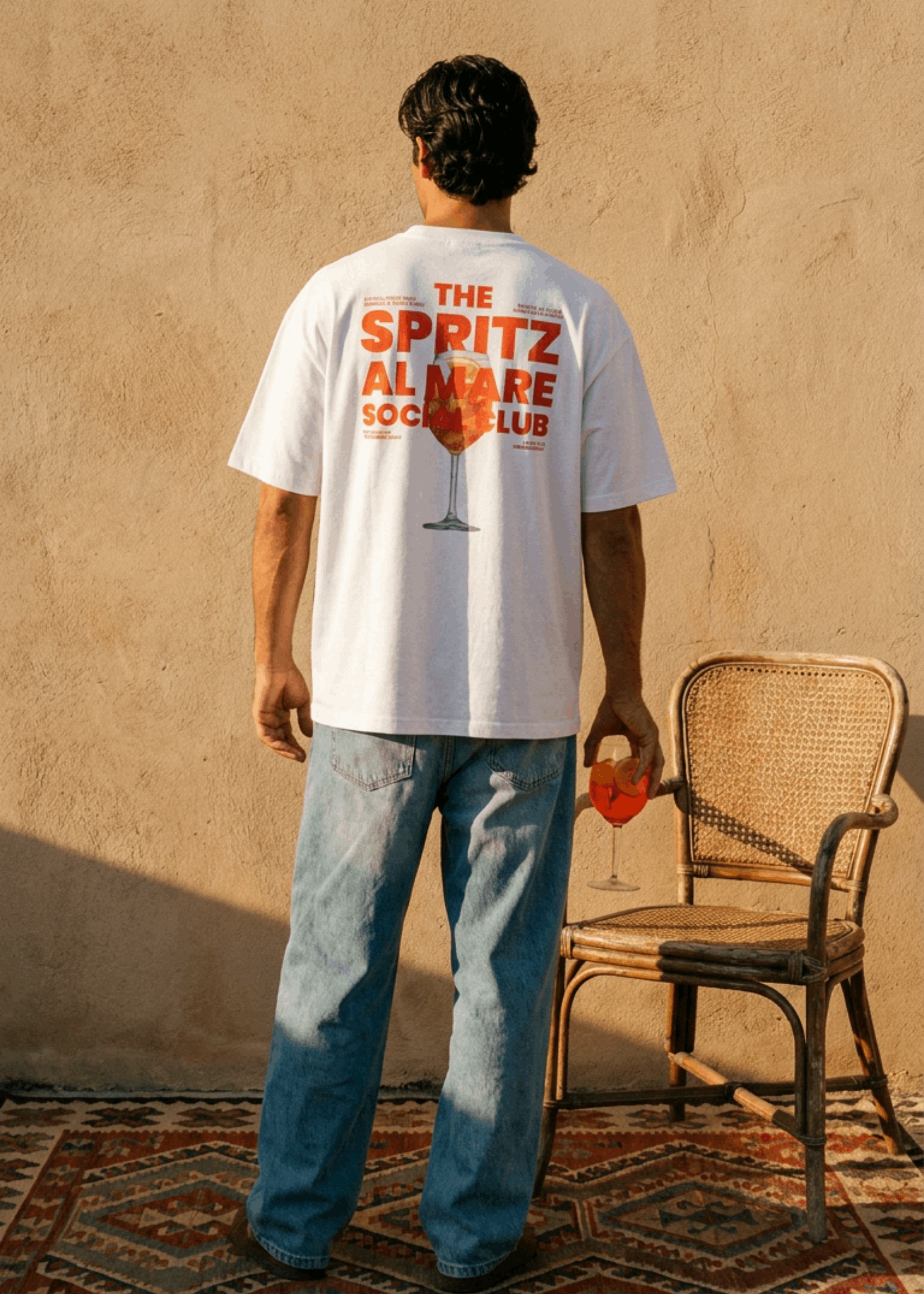 A man stands with his back to the camera wearing a white graphic t-shirt that reads 'THE SPRITZ ALMARE SOCIAL CLUB' with a red cocktail glass design, paired with blue jeans, in front of a textured beige wall next to a wicker chair.
