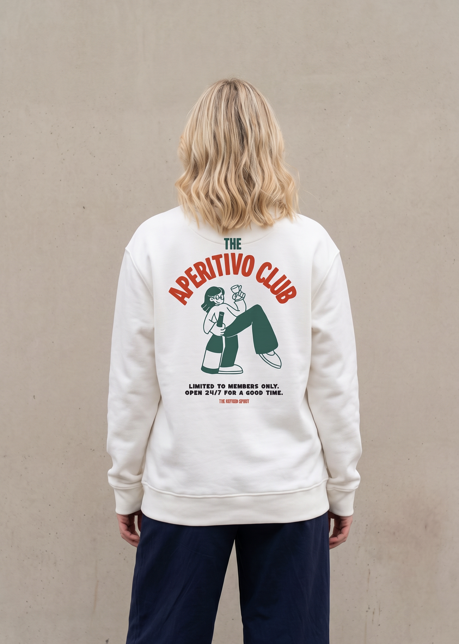 A woman viewed from behind wearing a cream-colored sweatshirt with a large graphic on the back that reads 'THE APETITIVO CLUB' in red and green, paired with dark navy pants, standing against a textured beige wall.