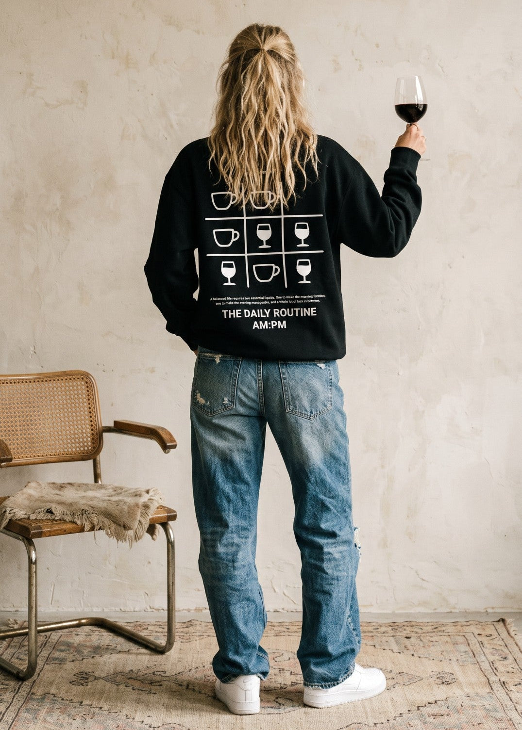 A woman stands with her back to the camera wearing a black sweatshirt with a white graphic print of coffee cups and the text 'THE DAILY ROUTINE 6AM-6PM'. She is wearing blue jeans and white sneakers, holding a glass of red wine in a room with a textured beige wall and a woven chair.