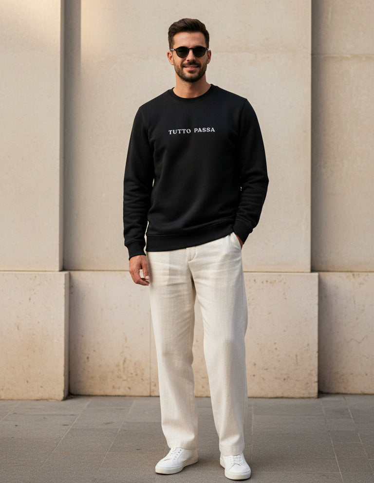 Man wearing a black sweatshirt with text and white pants standing against a beige wall.