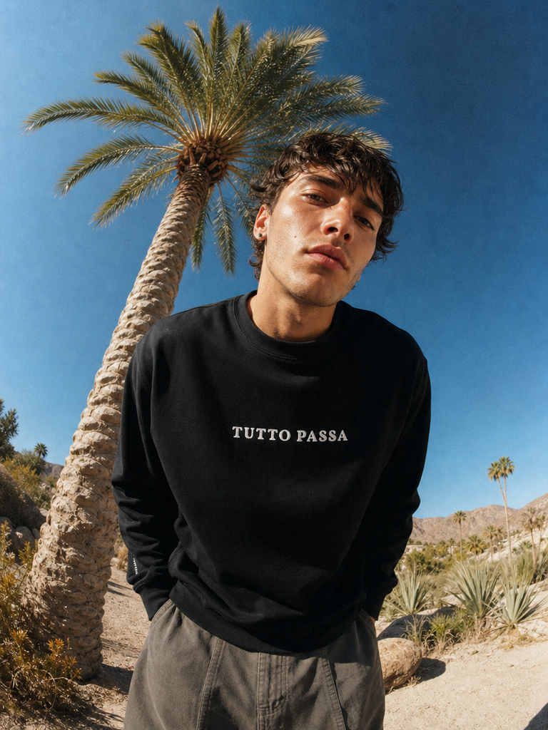 A young man wearing a black crewneck sweatshirt with 'TUTTO PASSA' printed in white across the chest, standing outdoors in a desert landscape with a palm tree and blue sky.