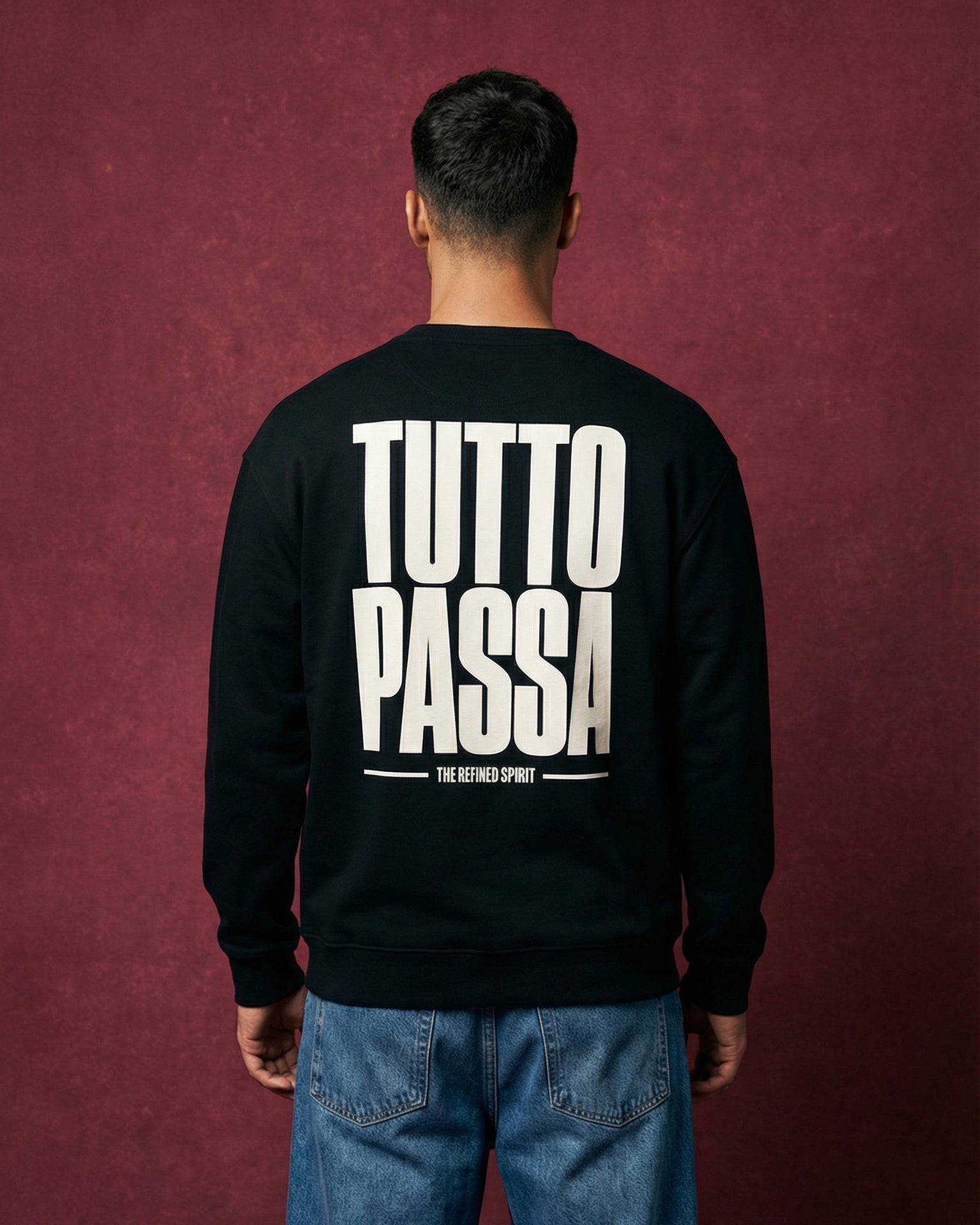 Person wearing a black sweatshirt with 'Tutto Passa' text on the back against a maroon background