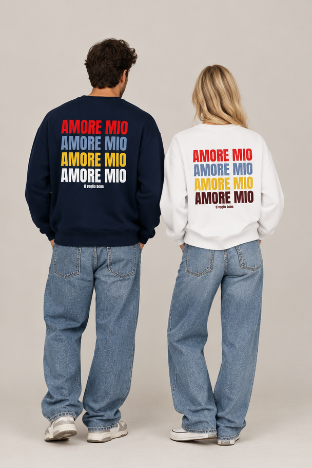 Two models from behind wearing matching crewneck sweatshirts with 'AMORE MIO' printed repeatedly in red, blue, yellow, and brown on the back. One sweatshirt is navy blue and the other is white, paired with light-wash jeans against a neutral studio background.