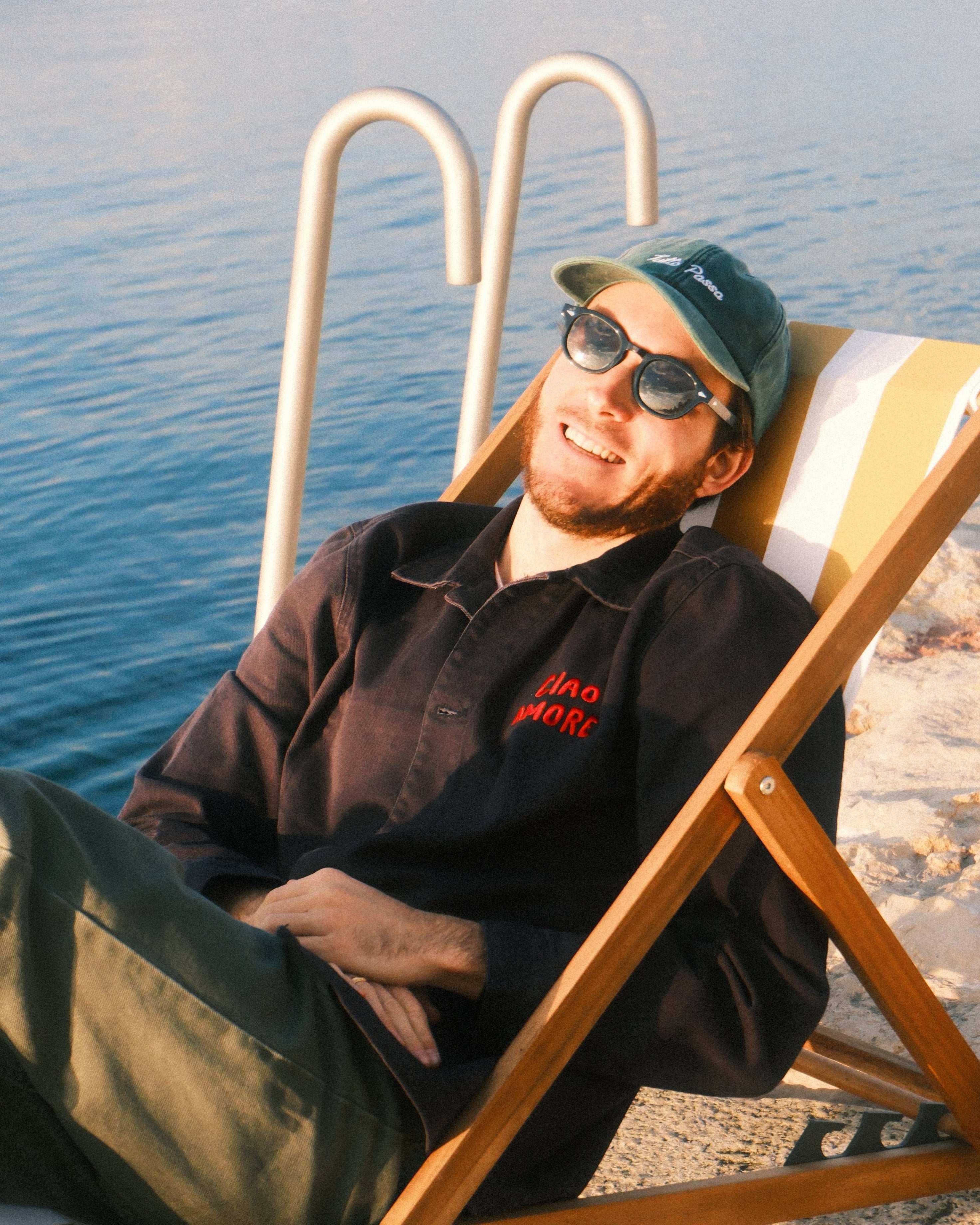 Person sitting on a wooden chair by a poolside wearing sunglasses and a cap.