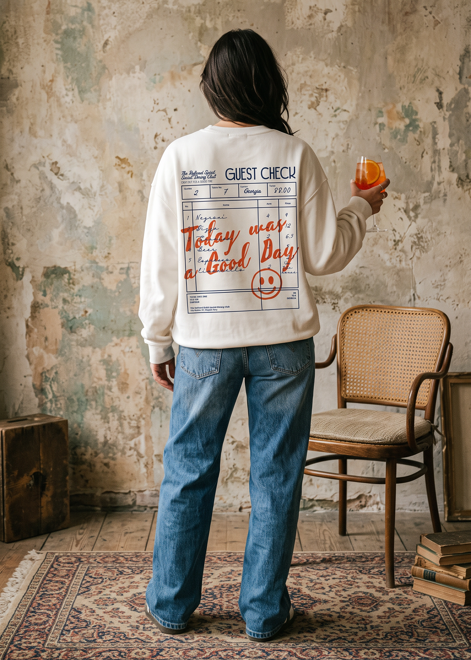 A person stands with their back to the camera wearing a cream-colored sweatshirt with a large graphic print on the back, paired with blue jeans. They are holding an orange drink in a rustic room with a distressed wall, a wooden chair, and a patterned rug.