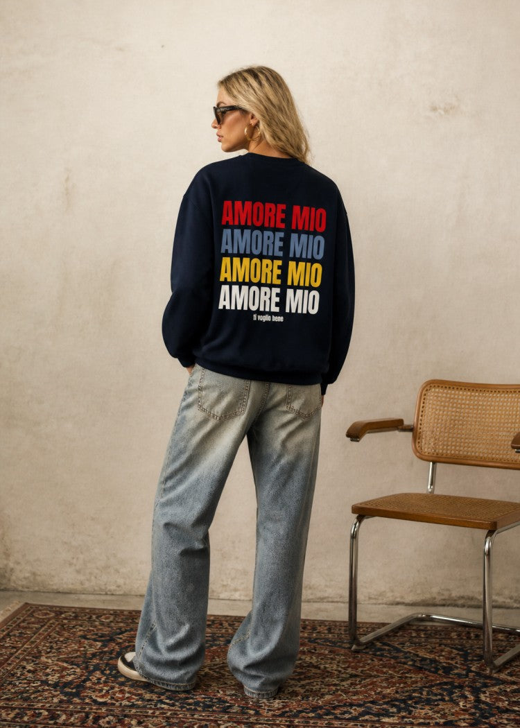 A woman stands with her back to the camera wearing a navy blue sweatshirt with 'AMORE Mio' printed in red, blue, yellow, and white on the back, paired with light-wash wide-leg jeans in a minimalist room with a textured wall and a wooden chair.