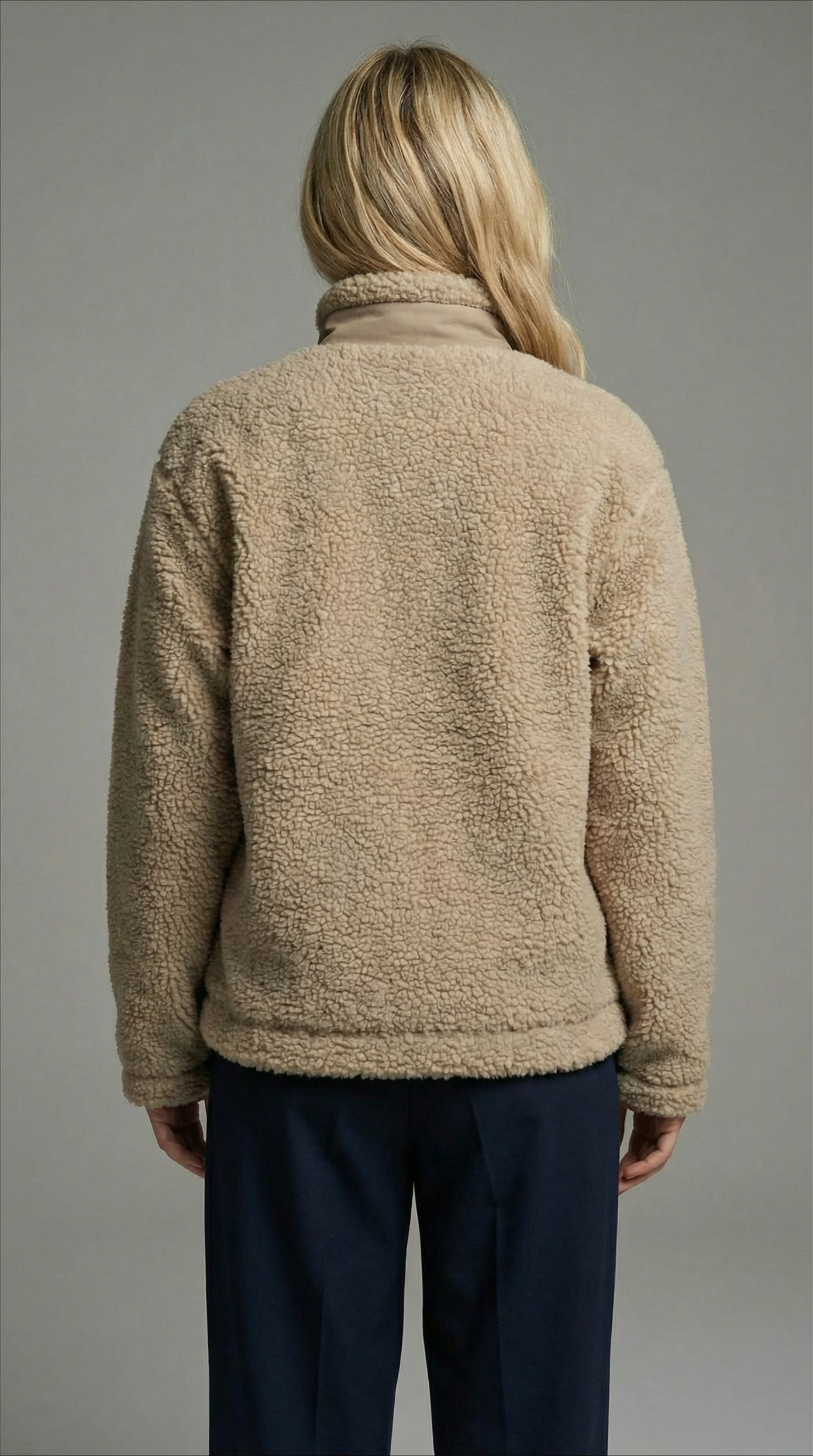 Person wearing a beige fleece jacket and dark pants against a gray background