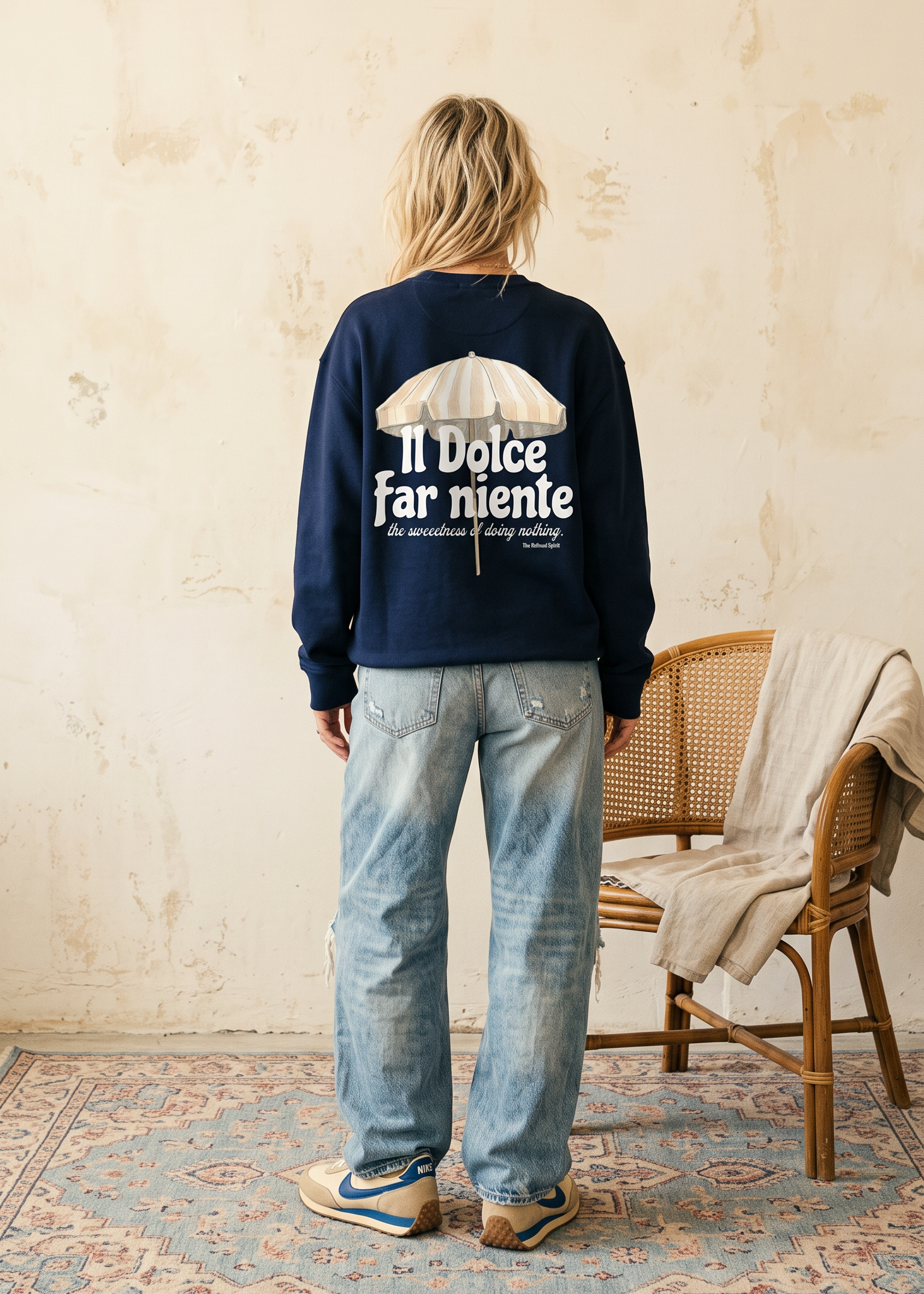 A woman stands with her back to the camera wearing a navy blue sweatshirt with a large white graphic and text on the back, paired with light-wash blue jeans. She is standing in a rustic room with a textured beige wall, a patterned rug, and a wooden chair with a beige throw blanket.