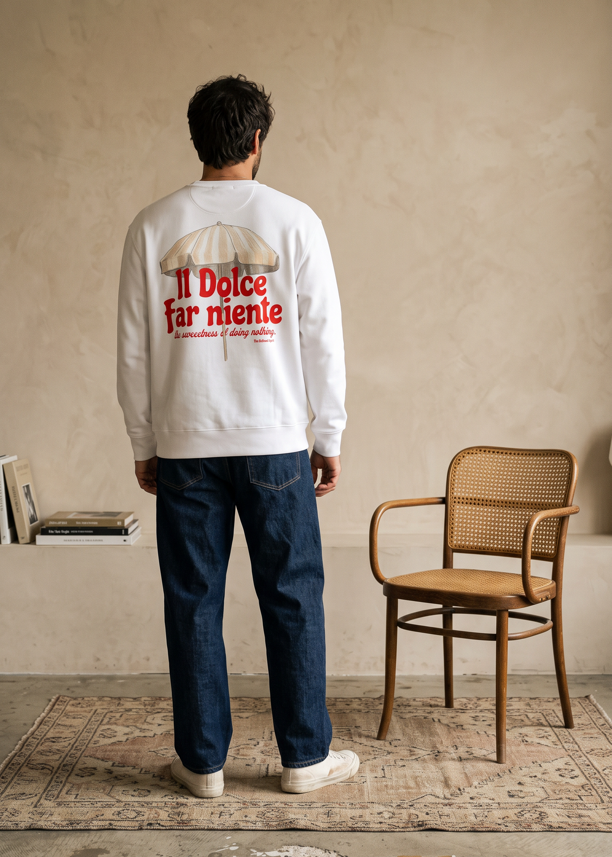 A man stands with his back to the camera wearing a white sweatshirt with a red and beige graphic on the back that reads 'Il Dolce far niente'. He is wearing blue jeans and white sneakers in a minimalist room with a textured beige wall, a wooden chair, and a patterned rug.