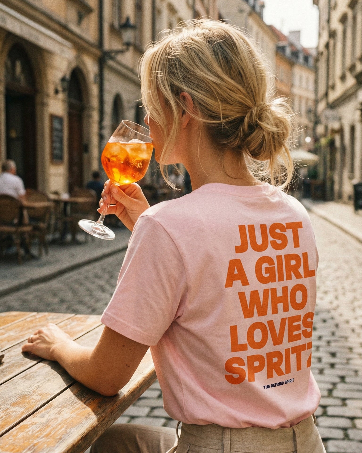 Woman sitting at an outdoor table holding a glass of orange juice, wearing a pink t-shirt with text.