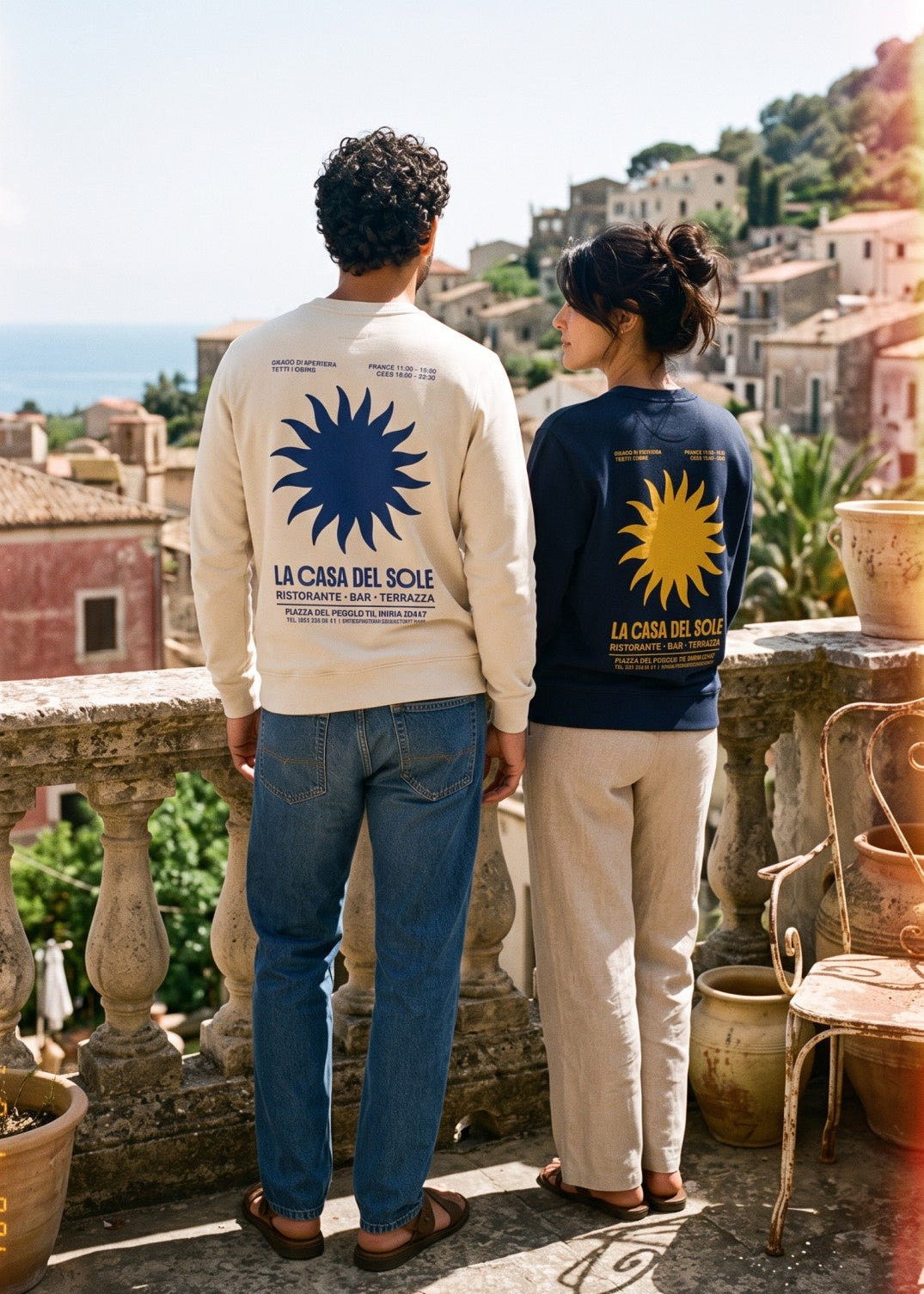 A man and a woman stand on a sunlit terrace overlooking a coastal town and sea, wearing matching oversized sweatshirts with large sun graphics and the text 'LA CASA DEL SOLE'.