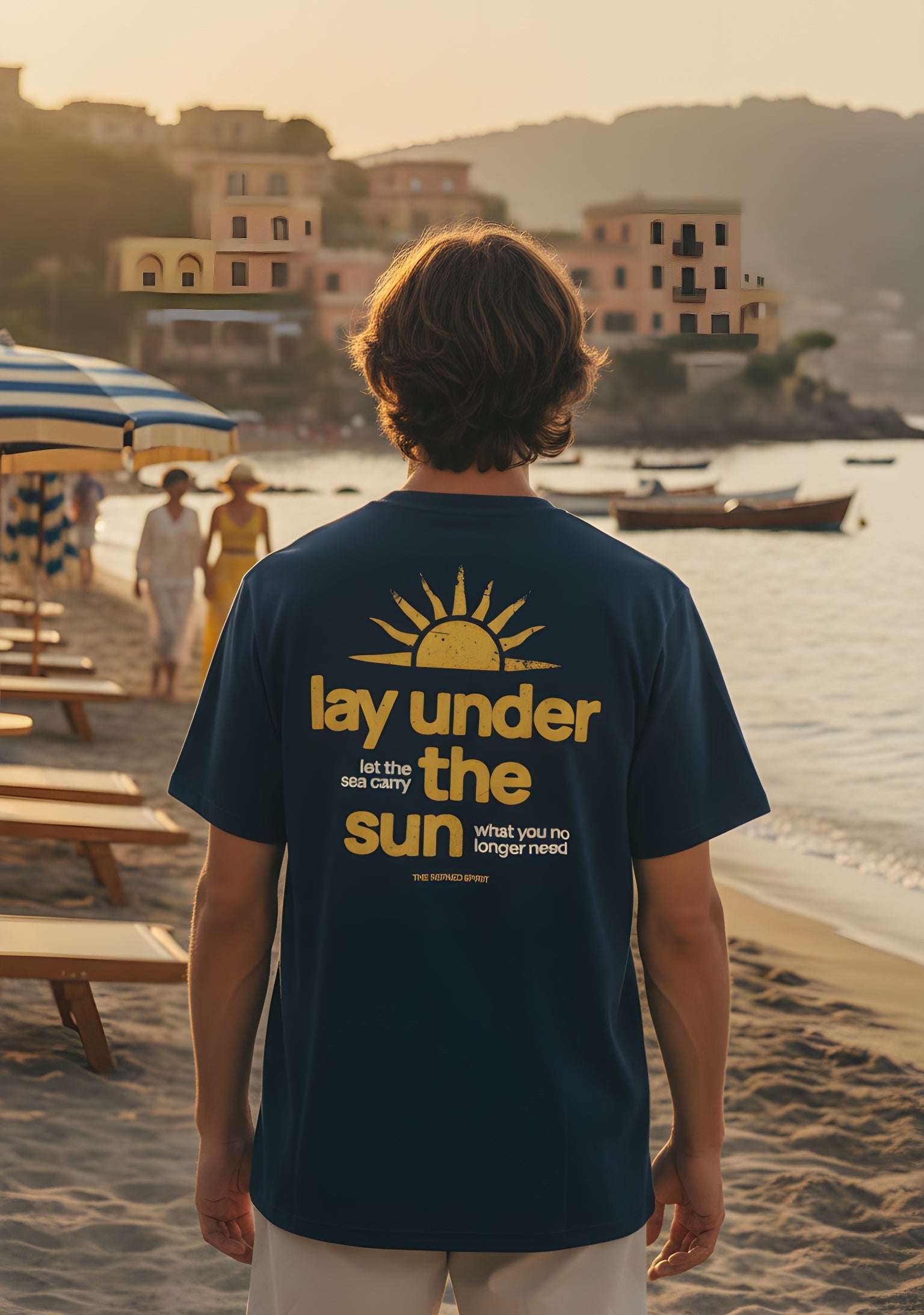 Person wearing a t-shirt with a beach-themed design on a sandy beach with buildings and water in the background.