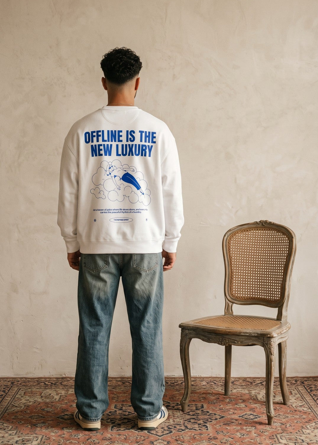 A man stands with his back to the camera wearing a white crewneck sweatshirt with a blue graphic print that reads 'OFFLINE IS THE NEW LUXURY'. He is wearing blue jeans and standing in a room with a textured beige wall, a vintage wooden cane-back chair, and a patterned rug.