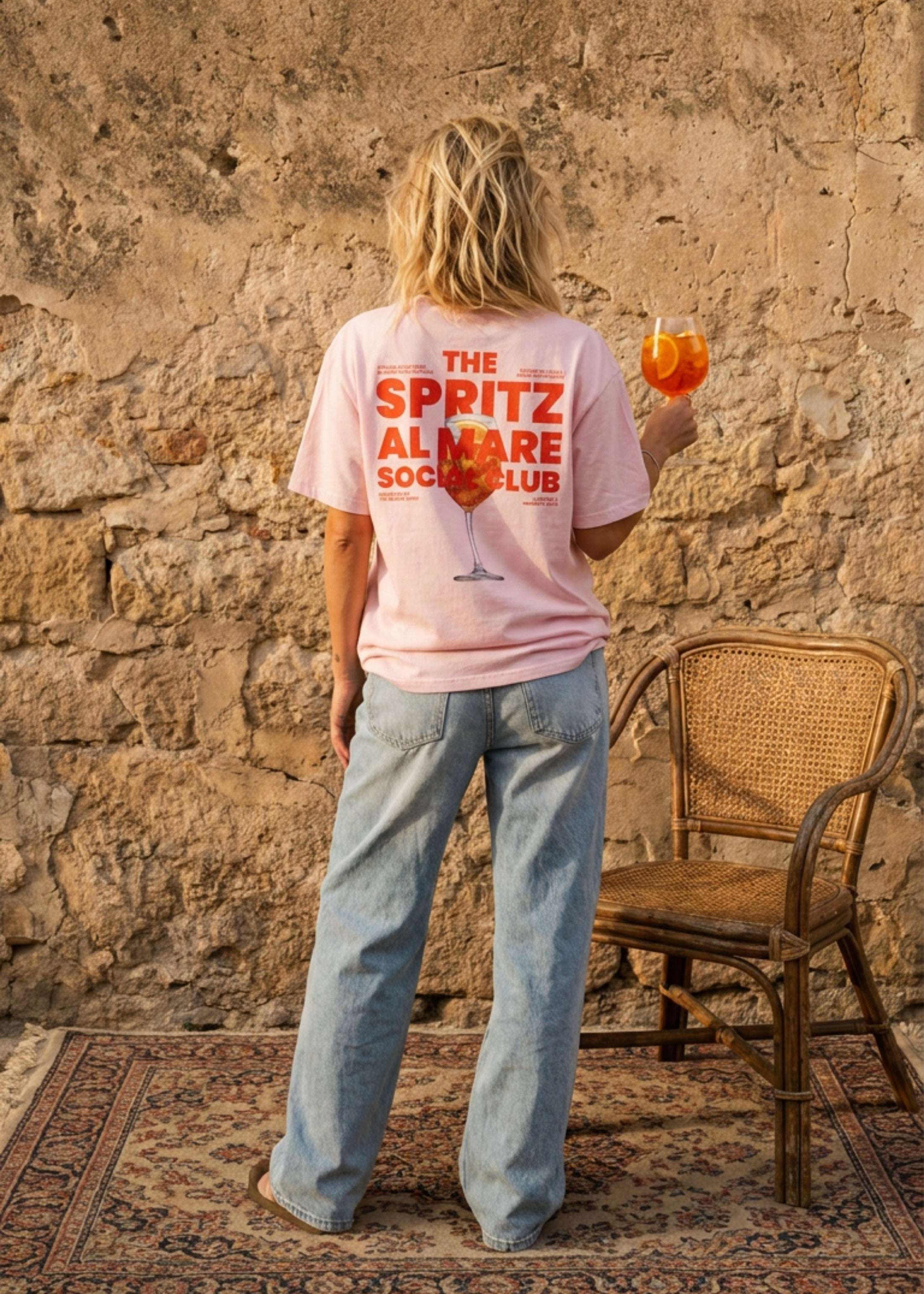 A woman stands with her back to the camera wearing a light pink t-shirt with orange text that reads 'THE SPRITZ AL MARE SOCIAL CLUB', paired with light blue jeans, holding an orange drink in front of a rustic stone wall.