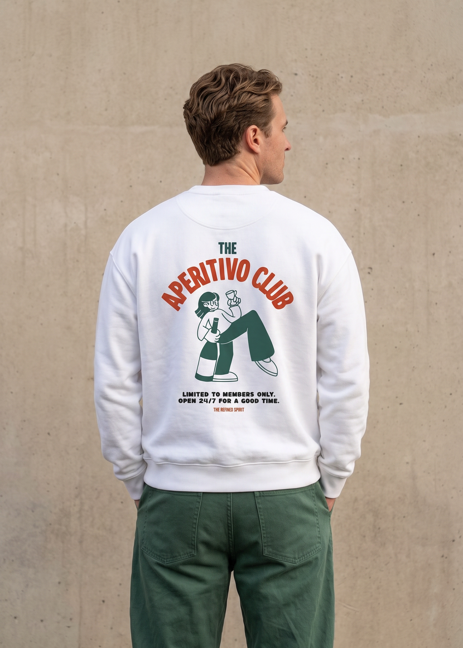A man viewed from behind wearing a white crewneck sweatshirt with a large graphic print on the back that reads 'THE APERITIVO CLUB' in red and green, paired with green pants, standing against a textured concrete wall.