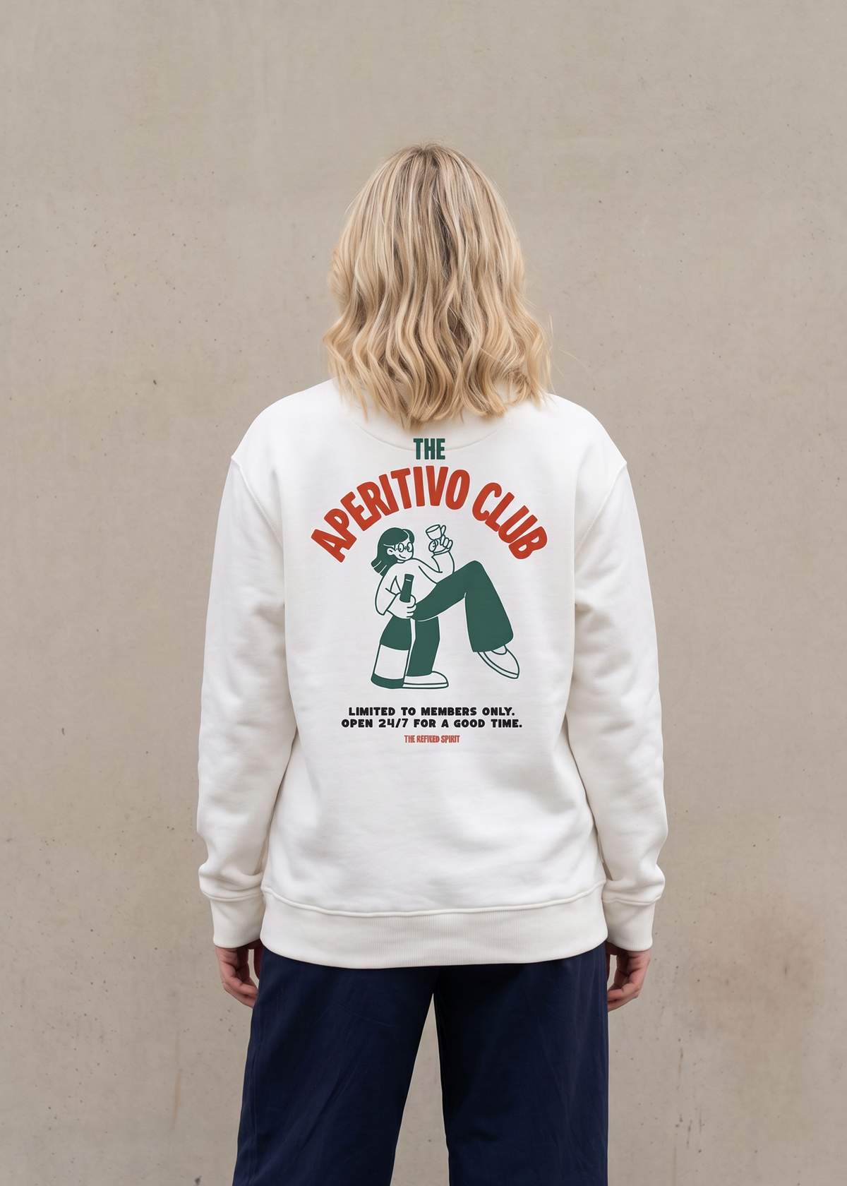 A woman viewed from behind wearing a cream-colored sweatshirt with a large graphic on the back that reads 'THE APETITIVO CLUB' in red and green, paired with dark navy pants, standing against a textured beige wall.