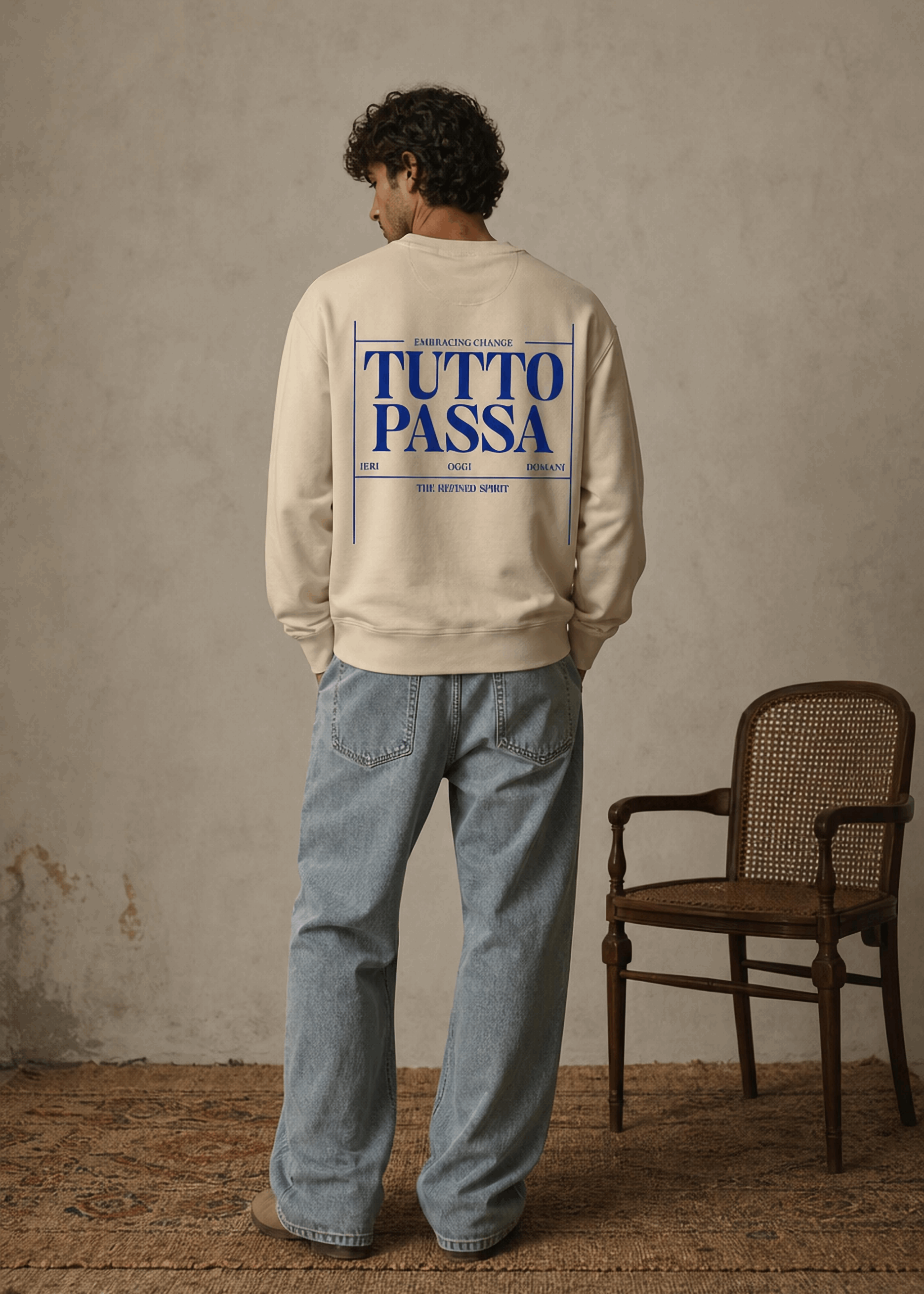 Person wearing a beige sweatshirt with 'TUTTO PASSA' text on the back, standing in a room with a chair.