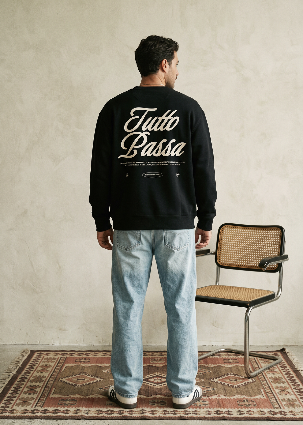 A man stands with his back to the camera wearing a black crewneck sweatshirt with 'Tutto Basta' printed in white script across the back, paired with light blue jeans. He is standing on a patterned rug next to a modern chair against a textured beige wall.