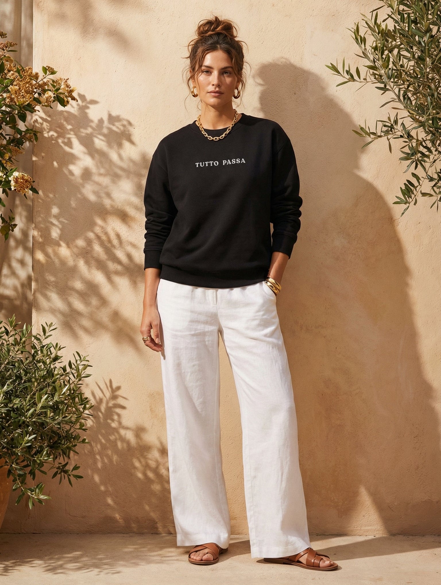 Woman wearing a black sweater and white pants standing against a beige wall with plants around.