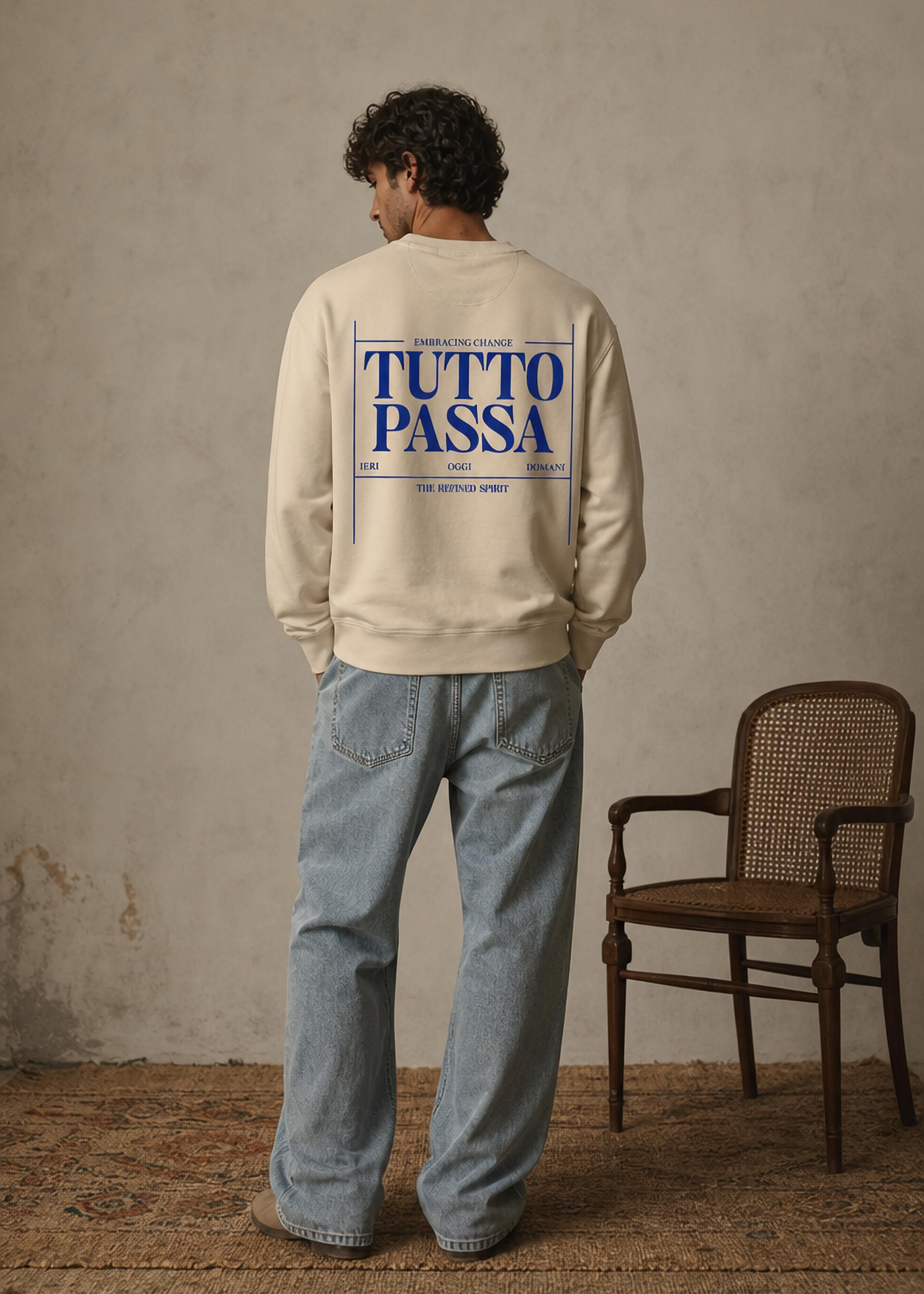 Person wearing a beige sweatshirt with 'TUTTO PASSA' text on the back, standing in a room with a chair.